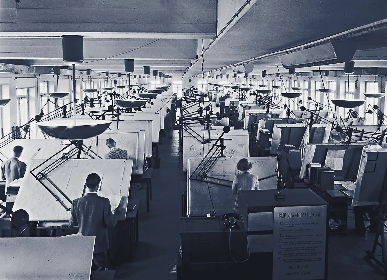 A drawing office in the 1950s: two long rows of draughtsmen at inclined drawing boards under anglepoise lamps and shaded pendants, the room running away to windows at the far end.